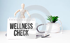 The text wellness check is written on notepad and wood man toy near a stethoscope on a blue background. Medical concept