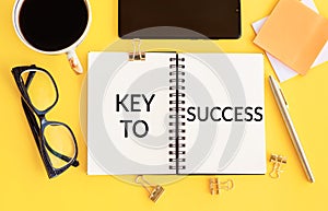 The text, the word Key To Success, is written in a notebook lying on a yellow table with a pen, coffee, glasses, sticks. Business