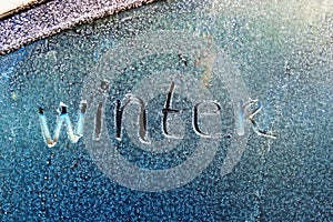 Text word winter scraped on frosty glass covered with white frost crystals on car blue window on winter cold clear morning