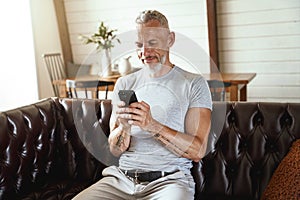 Texting message. Middle aged caucasian man in casual clothes using his smartphone while sitting on sofa in the living
