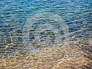 Texture of crystal clear light blue and turquoise transparent water with fine white sand base with bright reflection of sun in