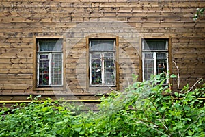 Texture of a wooden wall of an old house with three windows and frame covered with peeling white paint. In the windows are visible