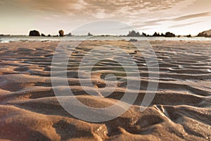 Texture of yellow sand waves in the beach with a cloudy sky and rocks in the background. Ideal for backgrounds or wallpapers