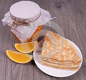 On a textured wooden background, a jar of orange syrup next to it is a plate of pancakes and sliced â€‹â€‹orange slices