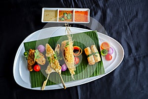 Thai appetizers spring rolls, crispy prawn, Thai sausage, chicken satay on black background.