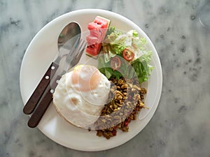 Thai food - spicy stirred fried pork chop served with rice, fried egg, and vegetables on a white dish on a white marble table