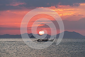 Thai longtail boat in the sea at sunset with big red sun and mountains silhouettes and clouds