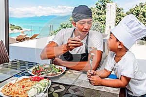 Thai man and his cute son having lunch at home. Playing game of cooks: apron, chef`s hat and thai food