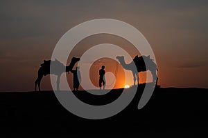 Thar desert, Rajasthan, India- 15.10.2019 : Silhouette of two cameleers and their camels at sand dunes. Cloud with setting sun,