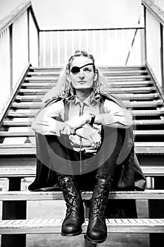 A theatrically dressed pseudo soldier or pirate with a blindfold in one eye sits on the stairs. Young man with a viking haircut