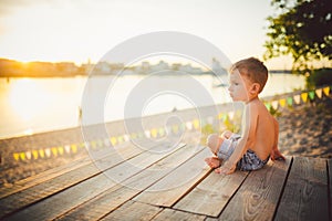 The theme is child and summer beach vacation. A small Caucasian boy sits sideways on a wooden pier and admires the view of the san