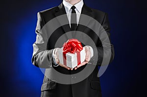 Theme holidays and gifts: a man in a black suit holds an exclusive gift in a white box wrapped with red ribbon and bow on a dark