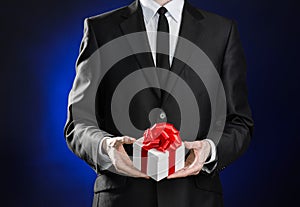 Theme holidays and gifts: a man in a black suit holds an exclusive gift in a white box wrapped with red ribbon and bow on a dark