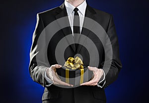 Theme holidays and gifts: a man in a black suit holds exclusive gift wrapped in a black box with gold ribbon and bow on a dark