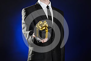 Theme holidays and gifts: a man in a black suit holds exclusive gift wrapped in a black box with gold ribbon and bow on a dark