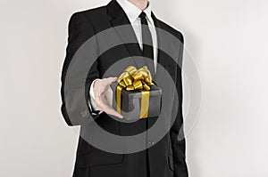 Theme holidays and gifts: a man in a black suit holds exclusive gift wrapped in a black box with gold ribbon and bow isolated on