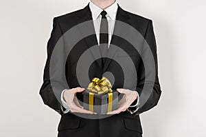 Theme holidays and gifts: a man in a black suit holds exclusive gift wrapped in a black box with gold ribbon and bow isolated on