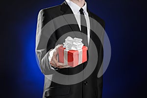 Theme holidays and gifts: a man in a black suit holds exclusive gift wrapped in red box with white ribbon and bow on a dark blue