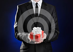 Theme holidays and gifts: a man in a black suit holds exclusive gift wrapped in red box with white ribbon and bow on a dark blue
