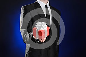 Theme holidays and gifts: a man in a black suit holds exclusive gift wrapped in red box with white ribbon and bow on a dark blue