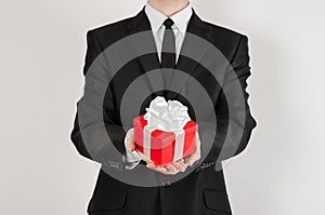 Theme holidays and gifts: a man in a black suit holds exclusive gift wrapped in red box with white ribbon and bow isolated on a