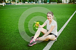 Theme sport and health. Beautiful young girl sitting resting on green grass, artificial turf stadium resting thirsty drink bottle