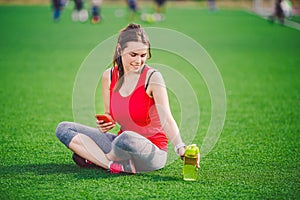 Theme sport health. beautiful young girl sitting resting on green grass. lawn stadium using technodogies. In handphone in ear