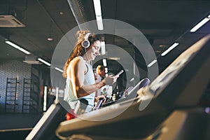Theme sport and music. A beautiful Caucasian woman running in gym on treadmill. On head big white headphones, the girl listens to