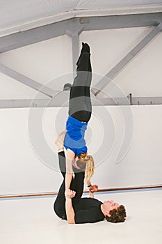 Theme is sports and acreage. A young Caucasian male and female couple practicing acrobatic yoga in a white gym on mats. a man lies