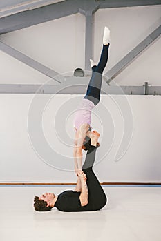 Theme is sports and acreage. A young Caucasian male and female couple practicing acrobatic yoga in a white gym on mats. a man lies