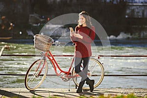 Theme tourism on bicycle and modern technology. Beautiful young caucasian woman stands near red retro bicycle on riverside river R