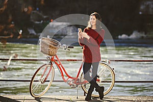 Theme tourism on bicycle and modern technology. Beautiful young caucasian woman stands near red retro bicycle on riverside river R