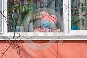 Therapeutic medical mask on a pink toy hare outside the window of the house. Theme stay at home during epidemics, protection from