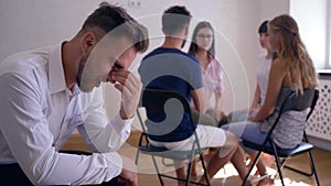 Therapy session, sad man thinks about problems on background of people sitting on chairs in circle