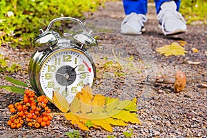 There is an alarm clock with autumn leaves and a mushroom on the path in the park. In the distance, the legs of a man in sneakers