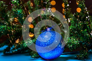 There is blue ball on the green xmas tree branch. There are glowing lights/bokeh on the background.