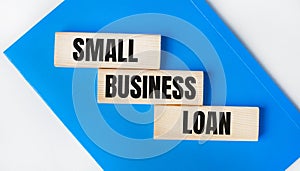 There is a blue notebook on a light gray background. Above are three wooden blocks with the words SMALL BUSINESS LOAN