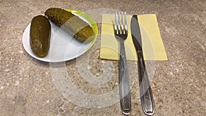 There is a ceramic saucer on the table with pickles on it. Next to it on a paper napkin is a metal knife and fork