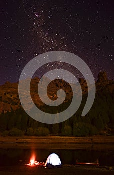 There is an illuminated tent near a river and a canoe on a starry night