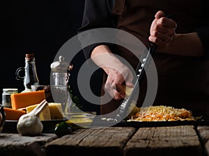 There are many products in the photo - garlic, cheese, sauce, spices. The chef grates cheese on a cutting board. Rough wooden