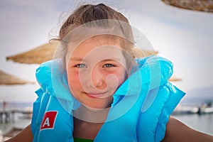 There is a portrait of a beautiful little girl in a life jacket. A happy child in a blue inflatable vest smiles cheerfully. Safe