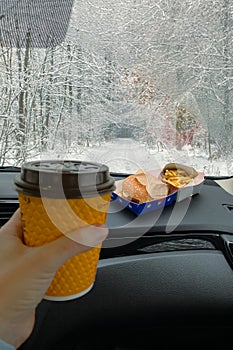 There's a hamburger, fries on the dashboard of the car. man is holding a cup of hot drink