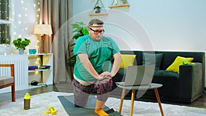 There s an obese man, wearing a green shirt and long socks stretching out doing lunges on the floor on a yoga mat, he is