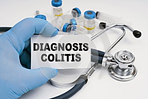 There is a stethoscope on the table, the doctor is holding a business card with the inscription - diagnosis COLITIS