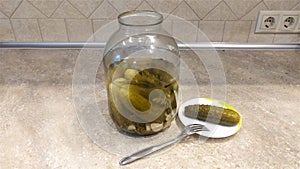 There is a three-liter glass jar with pickled cucumbers on the table. Next to the saucer, on which lies a cucumber and a fork. The