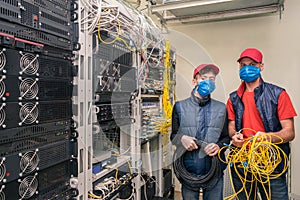 There are two technicians in medical masks in the server room. Specialists with a coil of wires in their hands work near the racks