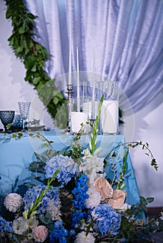 there are white candles on table with a blue tablecloth, a background from an arch with blue fabric, flowers are in front of the