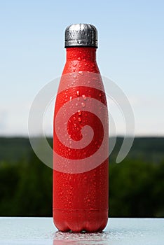 Thermo stainless bottle, sprayed with water. Sky and forest on background. On the glass desk. Thermos of matte red color.