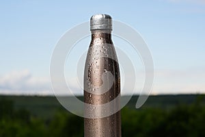 Thermo stainless bottle, sprayed with water. Sky and forest on background. Thermos of maro color.