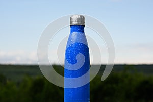Thermo stainless bottle, sprayed with water. Sky and forest on background. Thermos of matte blue color.